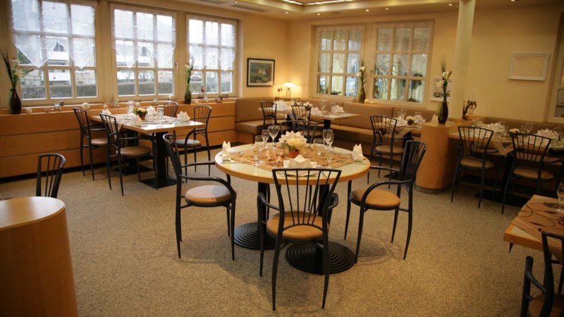 Ein elegantes Restaurant mit gedeckten Tischen, Stühlen und dekorativen Blumen. Große Fenster lassen viel Licht herein., © ateckhotel