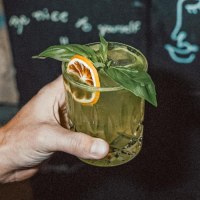 Eine tätowierte Hand hält ein grünes Getränk mit Basilikum und Orangenscheibe. Im Hintergrund ist eine Tafel mit Zeichnungen und Schrift zu sehen., © Bruno, Stuttgart Eine tätowierte Hand hält ein grünes Getränk mit Basilikum und Orangenscheibe. Im Hintergrund ist eine Tafel mit Zeichnungen und Schrift zu sehen., © Bruno, Stuttgart