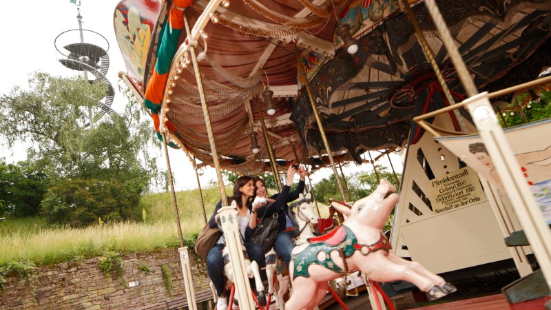 Zwei Personen fahren auf einem nostalgischen Karussell mit Pferdefiguren. Im Hintergrund ist ein spiralförmiger Aussichtsturm zu sehen., © SMG, Christoph Düpper Zwei Personen fahren auf einem nostalgischen Karussell mit Pferdefiguren. Im Hintergrund ist ein spiralförmiger Aussichtsturm zu sehen., © SMG, Christoph Düpper