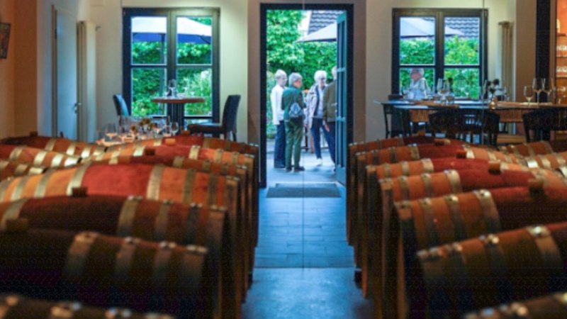 Barrique-F&auml;sser in einer Vinothek, im Hintergrund Menschen und Tische mit Gl&auml;sern, durch eine offene T&uuml;r ist ein gr&uuml;ner Garten sichtbar., &copy; Weingut W&ouml;hrwag