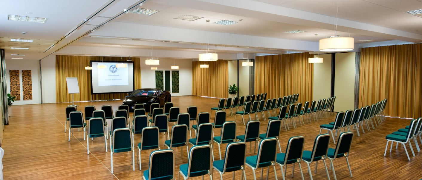 A conference room with rows of chairs, a car and a screen. The room has a wooden floor and yellow curtains., © Hotel Fortuna
