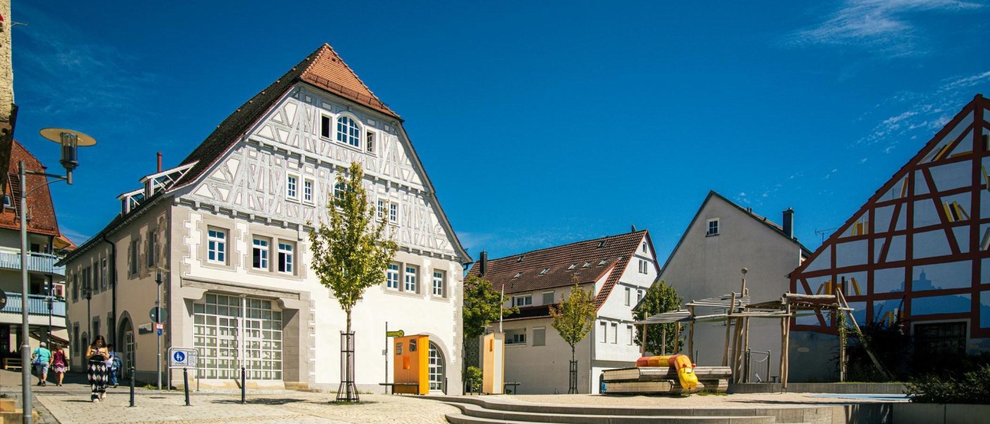 Historic half-timbered house on the market square of Vaihingen an der Enz, surrounded by modern buildings and a small playground, under a bright blue sky., © Stuttgart-Marketing GmbH, Sarah Schmid Historic half-timbered house on the market square of Vaihingen an der Enz, surrounded by modern buildings and a small playground, under a bright blue sky., © Stuttgart-Marketing GmbH, Sarah Schmid