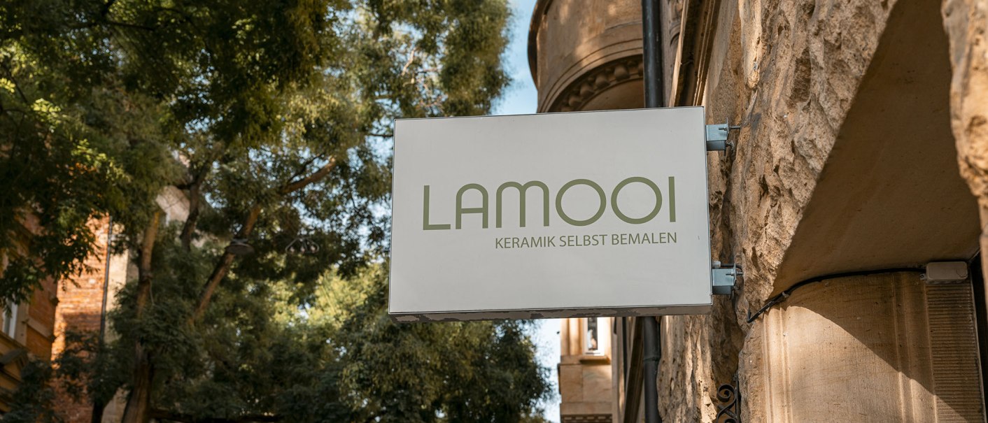Schild mit 'LAMOOI Keramik selbst bemalen' an einer Steinwand, umgeben von Bäumen., © SMG, Sarah Schmid