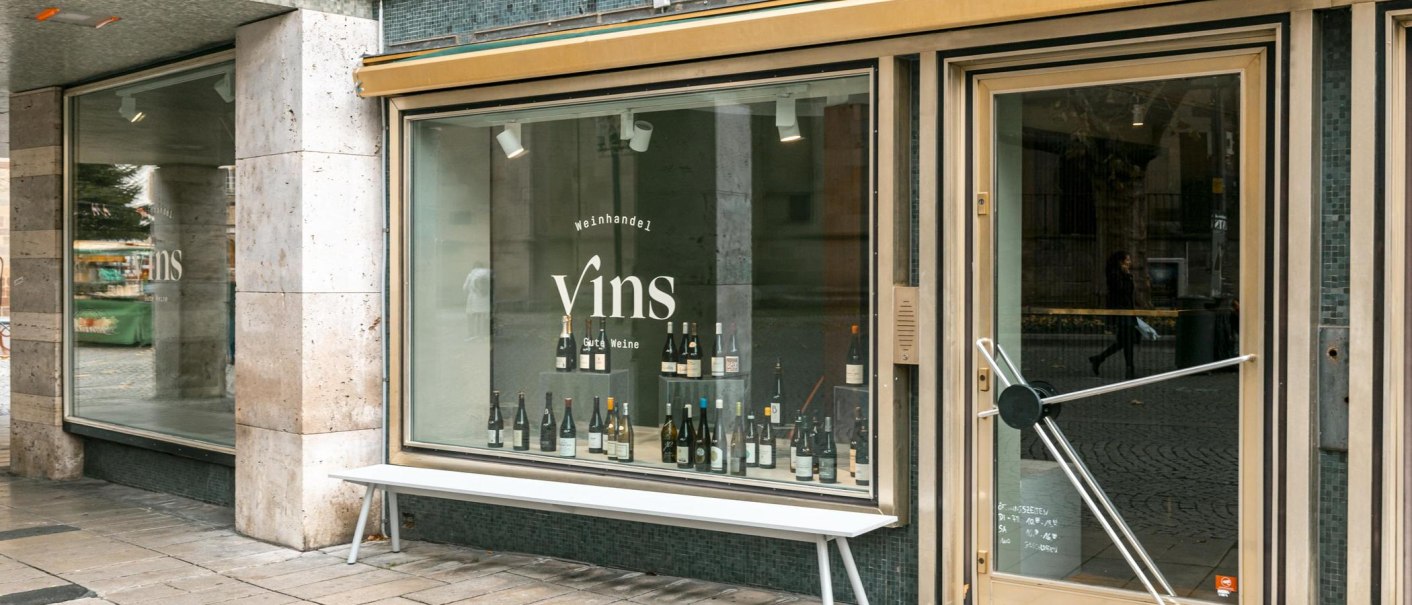 Schaufenster des Weinhandels 'Vins Gute Weine' mit Weinflaschen im Display und einer Bank davor., © SMG, Sarah Schmid