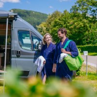 Ein Paar in blauen Badem&auml;nteln steht l&auml;chelnd vor einem modernen Wohnmobil auf einem sonnigen Campingplatz., &copy; Thomas Niederm&uuml;ller