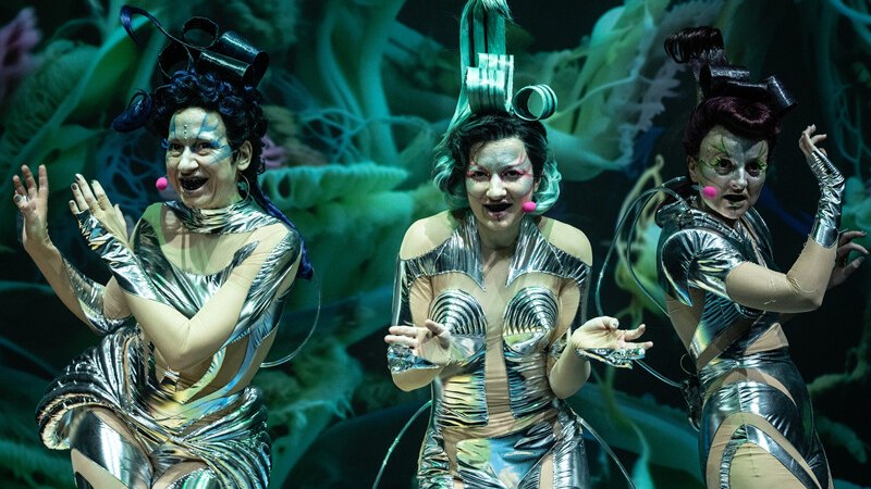 Three performers in silver, futuristic costumes perform on a stage in front of a green, abstract background., © Württembergische Staatstheater Stuttgart Three performers in silver, futuristic costumes perform on a stage in front of a green, abstract background., © Württembergische Staatstheater Stuttgart