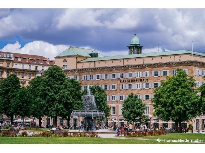 Ein belebter Platz in Stuttgart mit einem Brunnen und dem Gebäude "Carls Brauhaus" im Hintergrund, umgeben von Bäumen und Menschen., © Stuttgart Marketing GmbH Ein belebter Platz in Stuttgart mit einem Brunnen und dem Gebäude "Carls Brauhaus" im Hintergrund, umgeben von Bäumen und Menschen., © Stuttgart Marketing GmbH
