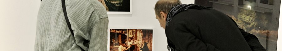 Zwei Personen betrachten aufmerksam Fotos an einer weißen Galeriewand. Die Bilder zeigen verschiedene Szenen, darunter Menschen und eine Schneelandschaft., © Palermo Galerie
