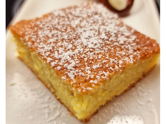 A juicy slice of lemon cake, sprinkled with powdered sugar, lies on a plate. A dollop of cream can be seen in the background., © Kultweine-Kochwelten-Kunstücke - Kochschule Sonja Lenz A juicy slice of lemon cake, sprinkled with powdered sugar, lies on a plate. A dollop of cream can be seen in the background., © Kultweine-Kochwelten-Kunstücke - Kochschule Sonja Lenz
