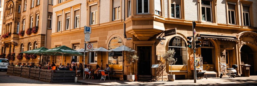 Street café with parasols in front of a historic building. People are sitting outside, enjoying the weather. Traffic signs and a Vespa are visible., © SMG Stuttgart Marketing GmbH - Sarah Schmid