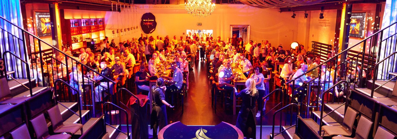 Veranstaltungsraum mit G&auml;sten an Tischen, warm beleuchtet, Kronleuchter und B&uuml;hne. Schriftzug 'AFTER-WORK-MAGIC' und 'STROTMANNS' sichtbar., &copy; Strotmanns Magic Lounge