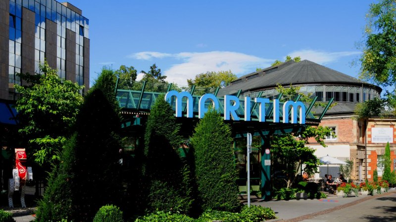 Außenansicht des Maritim Hotels mit markantem Glasdach und umgeben von Bäumen und Pflanzen. Ein Weg führt am Gebäude entlang., © Maritim Hotelgesellschaft mbH Außenansicht des Maritim Hotels mit markantem Glasdach und umgeben von Bäumen und Pflanzen. Ein Weg führt am Gebäude entlang., © Maritim Hotelgesellschaft mbH