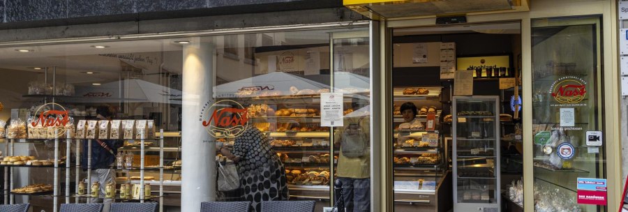 Exterior view of Cafe Nast with large windows through which baked goods and customers can be seen. A yellow sign with the name of the café hangs above the entrance., © SMG, Sarah Schmid