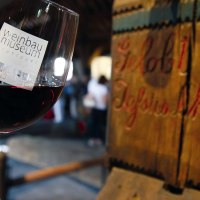 Ein Glas Rotwein mit dem Logo des Weinbaumuseums Stuttgart, im Hintergrund eine alte Holzpresse mit der Inschrift 'Gelobt sei Jesus Christus'., © Stuttgart-Marketing GmbH