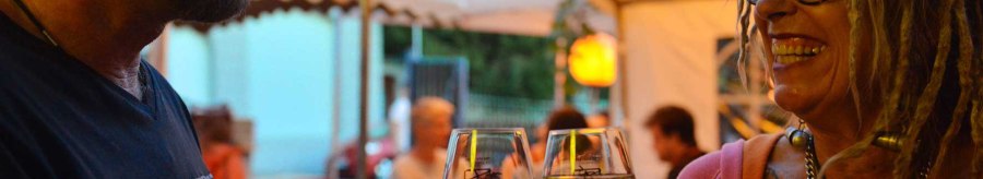 Two smiling people clink glasses of wine under a tent at a summer event., © Weingut Ruoff Two smiling people clink glasses of wine under a tent at a summer event., © Weingut Ruoff