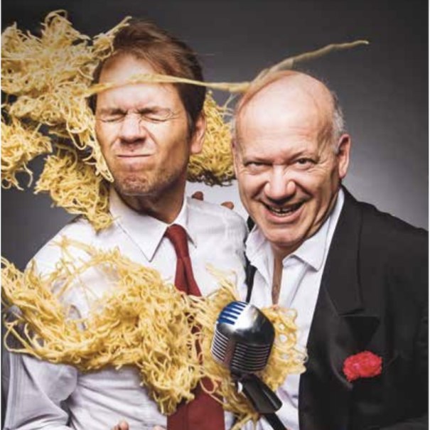 Two men in suits, one with noodles on his face, the other holding a microphone. Both are smiling and appear humorous., © Renitenztheater Stuttgart e.V. Two men in suits, one with noodles on his face, the other holding a microphone. Both are smiling and appear humorous., © Renitenztheater Stuttgart e.V.