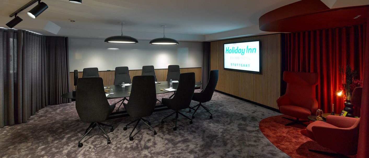 Moderner Konferenzraum mit gro&szlig;em Tisch, schwarzen St&uuml;hlen und rotem Sitzbereich. An der Wand ein Holiday Inn Stuttgart Schild., &copy; Holiday Inn Stuttgart