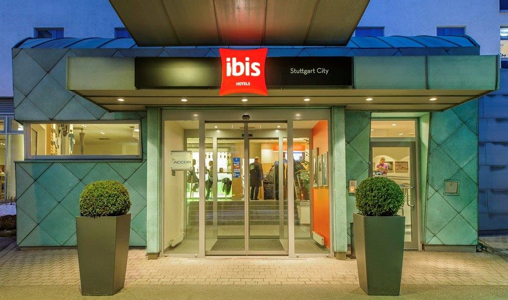 Eingang des Ibis Hotels Stuttgart City mit beleuchtetem Schild und Glasdrehtür, flankiert von zwei Pflanzen in modernen Pflanzgefäßen., © TOMAS Eingang des Ibis Hotels Stuttgart City mit beleuchtetem Schild und Glasdrehtür, flankiert von zwei Pflanzen in modernen Pflanzgefäßen., © TOMAS