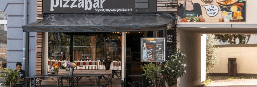 Außenansicht einer Pizzabar mit Terrasse, Blumen und einem Werbeplakat. Ein Mann sitzt an einem Tisch. Die Markise ist schwarz., © Stuttgart Marketing GmbH, Sarah Schmid Außenansicht einer Pizzabar mit Terrasse, Blumen und einem Werbeplakat. Ein Mann sitzt an einem Tisch. Die Markise ist schwarz., © Stuttgart Marketing GmbH, Sarah Schmid
