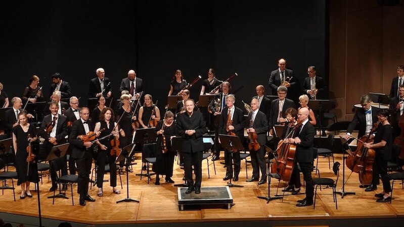 Das Stuttgarter Ärzteorchester e.V. steht auf einer Bühne. Musiker mit verschiedenen Instrumenten, ein Dirigent in der Mitte., © Stuttgarter Ärzteorchester e.V.