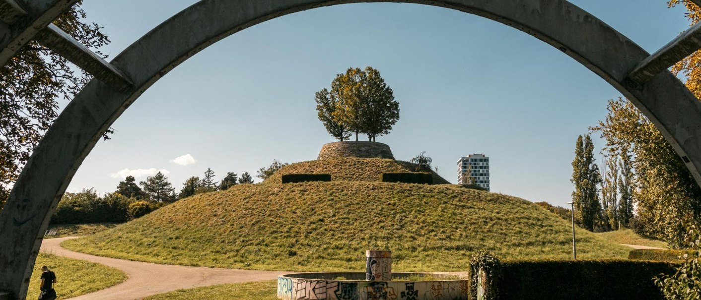 Grasbewachsener Hügel mit Baum, eingerahmt von Betonbogen. Im Hintergrund modernes Gebäude und blauer Himmel., © Stuttgart Marketing GmbH, Sarah Schmid