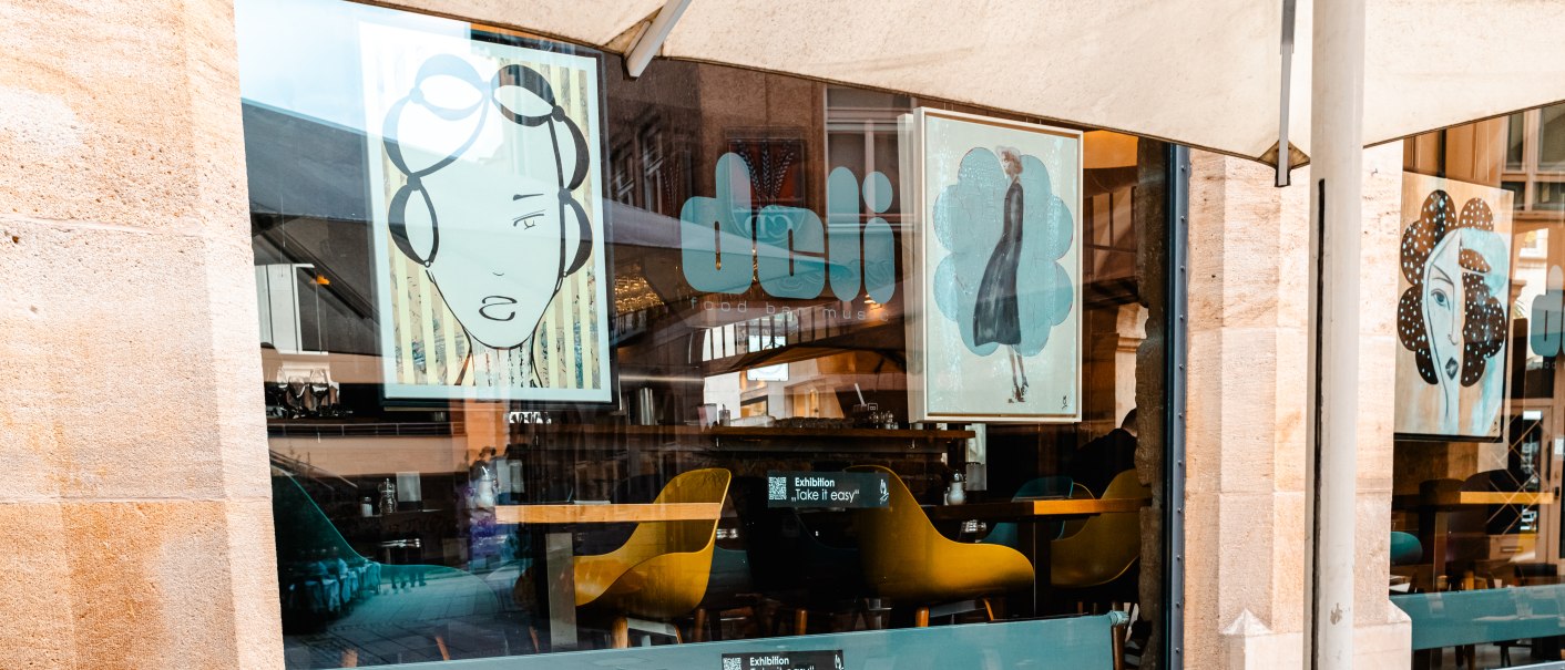 Ein Café namens 'Deli' mit großen Fenstern, dekoriert mit stilisierten Kunstwerken. Gelbe Stühle sind im Inneren sichtbar., © SMG Stuttgart Marketing GmbH - Sarah Schmid