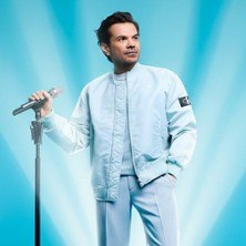 Eine Person in einem hellblauen Outfit posiert mit einem Mikrofon vor einem strahlend blauen Hintergrund., © links im Bild Eine Person in einem hellblauen Outfit posiert mit einem Mikrofon vor einem strahlend blauen Hintergrund., © links im Bild