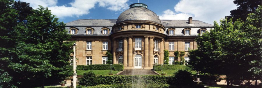 Die Villa Reitzenstein mit imposanter Fassade, einem Brunnen im Vordergrund und einer Flagge auf dem Dach, umgeben von Bäumen., © Staatsministerium Baden-Württemberg