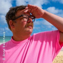 Person in pinkem T-Shirt mit Brille und Schnurrbart schaut in die Ferne am Strand. Der Himmel ist blau mit einigen Wolken., © links im Bild Person in pinkem T-Shirt mit Brille und Schnurrbart schaut in die Ferne am Strand. Der Himmel ist blau mit einigen Wolken., © links im Bild