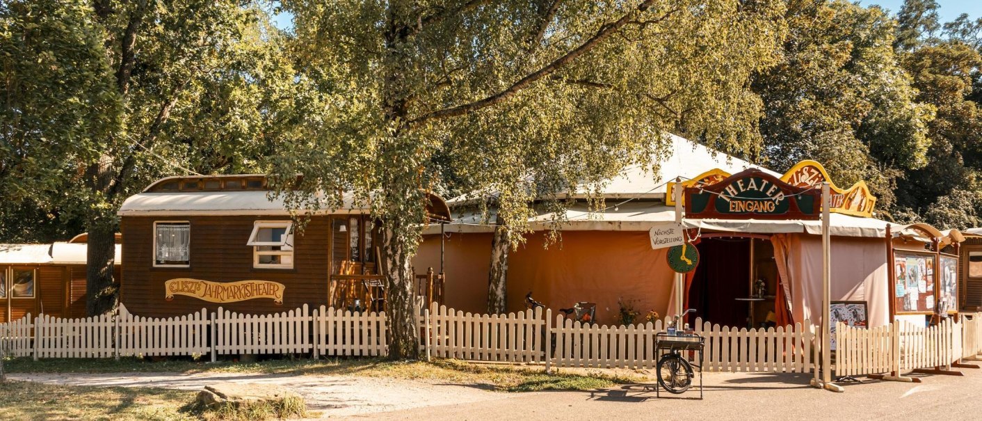 Eingang zu einem Jahrmarkttheater mit Holzzaun, Bäumen und einem Schild für die nächste Vorstellung. Ein Fahrrad steht daneben., © Stuttgart Marketing GmbH, Sarah Schmid