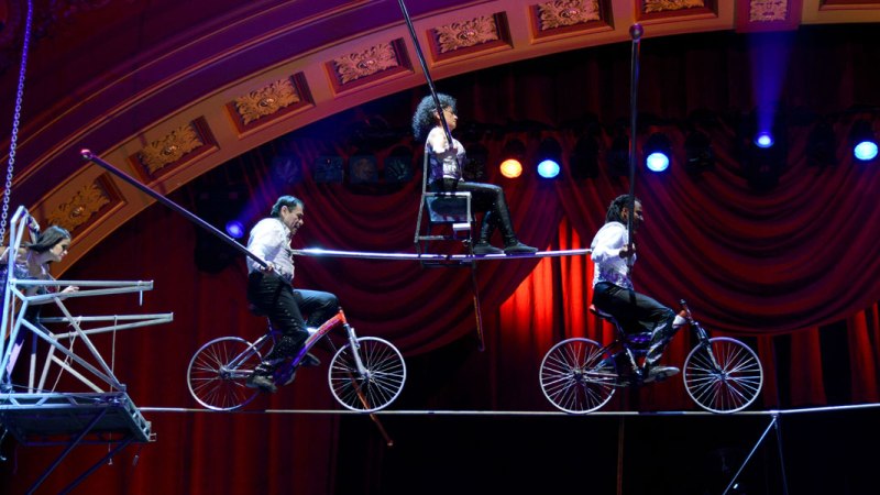 Artisten balancieren auf Fahrrädern und einem Stuhl auf einem Hochseil im Zirkus, umgeben von rotem Vorhang und bunten Lichtern., © Thierry Bissat