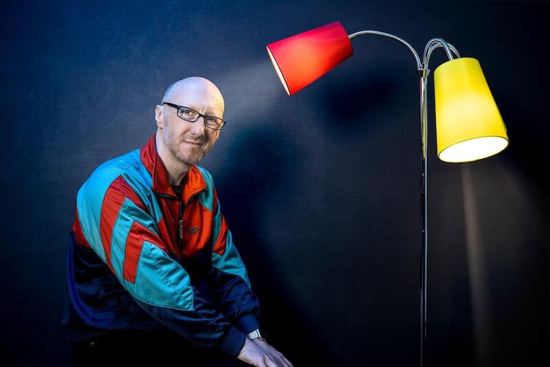 A man in a colorful jacket sits next to a floor lamp with red and yellow shades in front of a dark background., © Rosenau Kultur e.V. A man in a colorful jacket sits next to a floor lamp with red and yellow shades in front of a dark background., © Rosenau Kultur e.V.