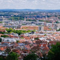 Panoramablick auf eine Stadt mit roten Dächern und modernen Gebäuden, umgeben von grünen Hügeln und bewölktem Himmel., © Thomas Niedermüller Panoramablick auf eine Stadt mit roten Dächern und modernen Gebäuden, umgeben von grünen Hügeln und bewölktem Himmel., © Thomas Niedermüller