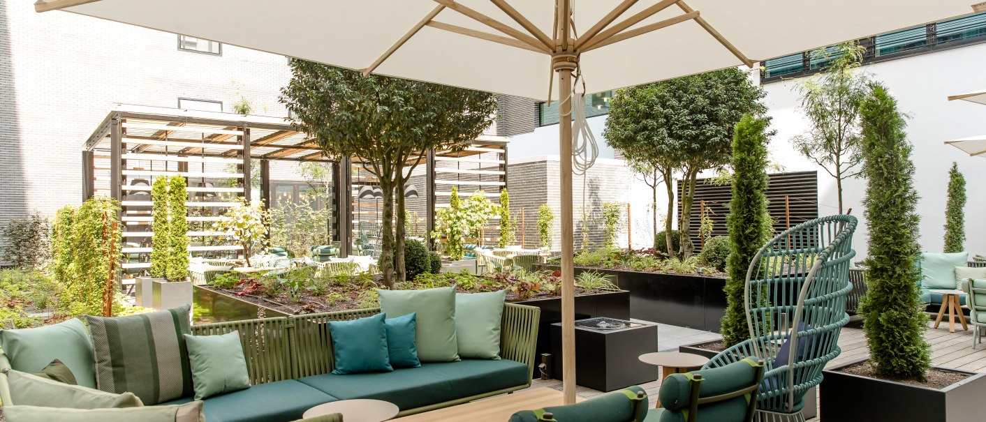 Moderne Terrasse mit grünen Sitzmöbeln, großem Sonnenschirm und üppiger Bepflanzung. Einladende Atmosphäre mit Holzboden und stilvollen Details., © Motel One - Stuttgart Hauptbahnhof