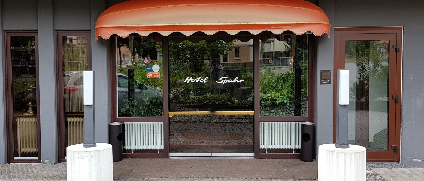 The entrance to the Hotel Spahr, featuring a glass facade, an orange canopy, and plants in the background. Two white columns stand in front of the door., &copy; Hotel Spahr
