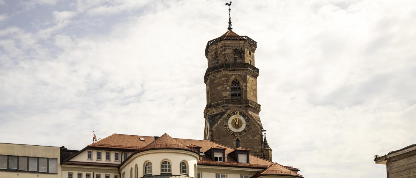 Tower of the collegiate church in Stuttgart with clock and weathercock, surrounded by buildings under a cloudy sky., &copy; SMG Stuttgart Marketing GmbH - Sarah Schmid