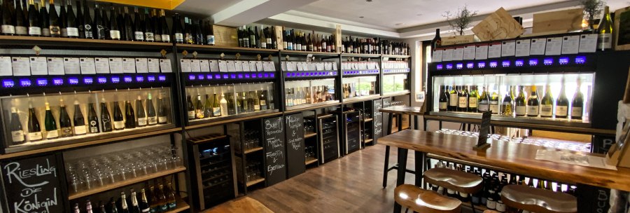 Weinladen mit Regalen voller Weinflaschen, Verkostungstischen und Gl&auml;sern. Moderne Einrichtung mit Holz- und Metallelementen., &copy; Sitt-Wein