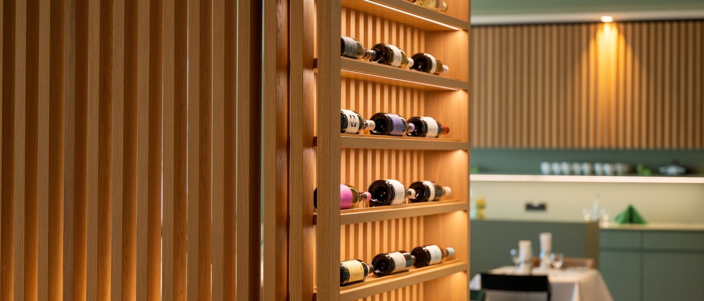 Ein Weinregal aus Holz in einem modernen Restaurant. Im Hintergrund sind gedeckte Tische und eine grüne Wand zu sehen., © Erikson Hotel Ein Weinregal aus Holz in einem modernen Restaurant. Im Hintergrund sind gedeckte Tische und eine grüne Wand zu sehen., © Erikson Hotel