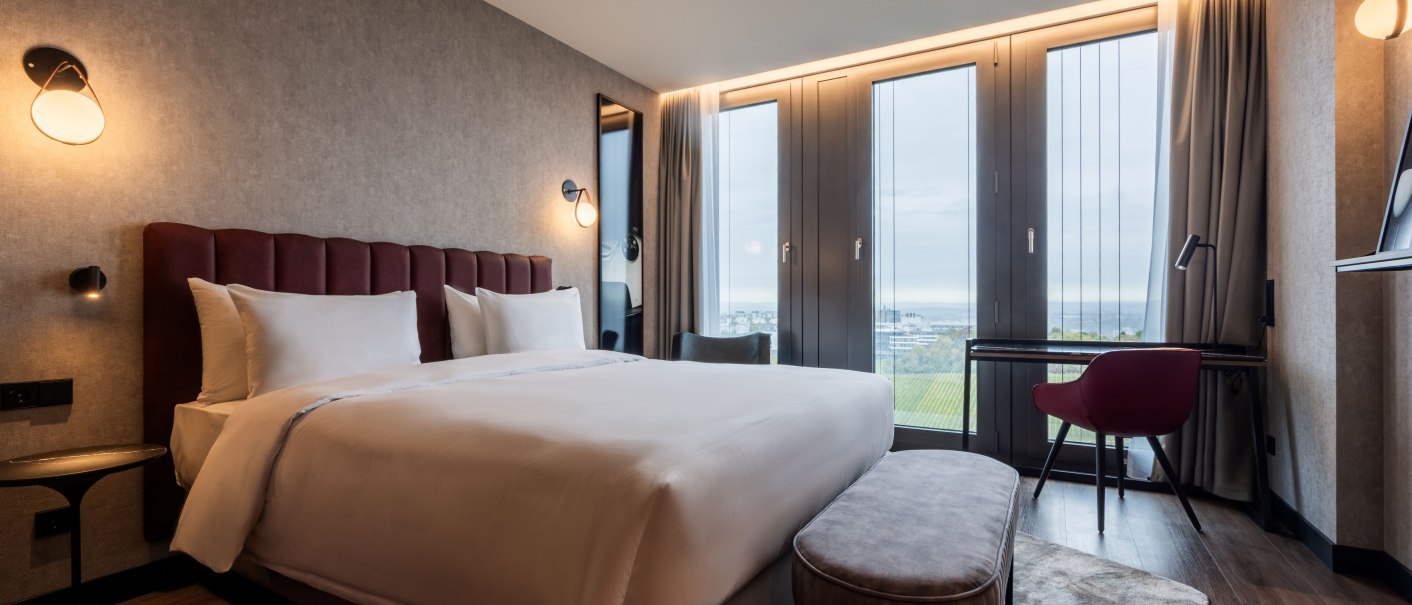 Modernes Hotelzimmer mit gro&szlig;em Bett, Schreibtisch und Blick auf Weinberge durch gro&szlig;e Fenster. Elegante Beleuchtung und stilvolle Einrichtung., &copy; Radisson Hotel Group