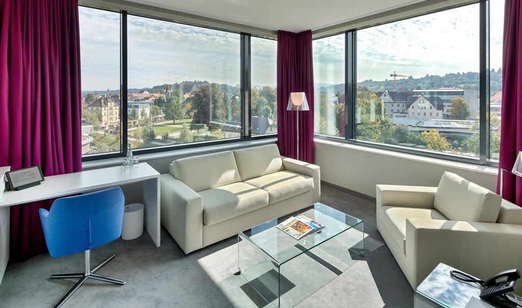 Modern suite with large windows, white sofas, glass table, desk and purple curtains. View of a cityscape., © Hotel am Remspark Modern suite with large windows, white sofas, glass table, desk and purple curtains. View of a cityscape., © Hotel am Remspark