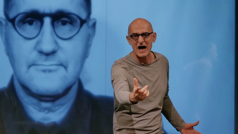 Ein Mann gestikuliert leidenschaftlich vor einem großen Porträt von Bertolt Brecht., © tri-bühne e.V. Ein Mann gestikuliert leidenschaftlich vor einem großen Porträt von Bertolt Brecht., © tri-bühne e.V.