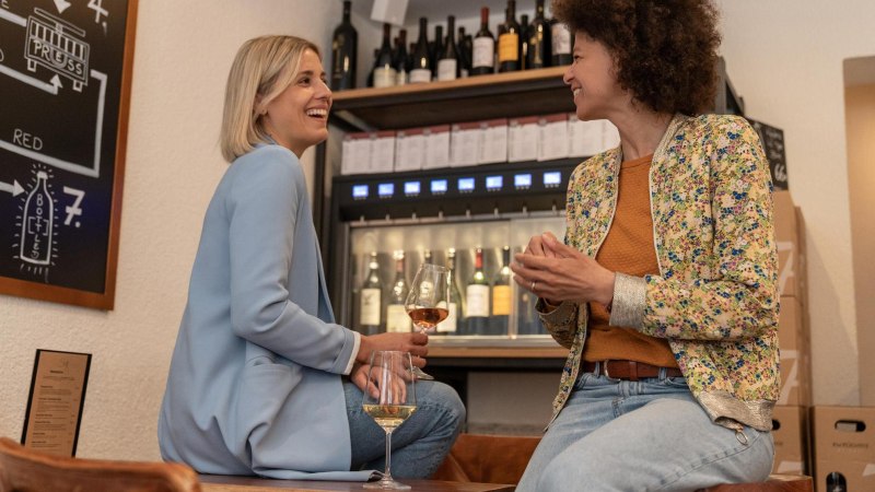 Two women are sitting on high stools, one of them is holding a glas of wine. They are laughing at each other. In the background is a wine dispenser with wine bottles that can be tapped., © Stuttgart-Marketing GmbH, Martina Denker Two women are sitting on high stools, one of them is holding a glas of wine. They are laughing at each other. In the background is a wine dispenser with wine bottles that can be tapped., © Stuttgart-Marketing GmbH, Martina Denker