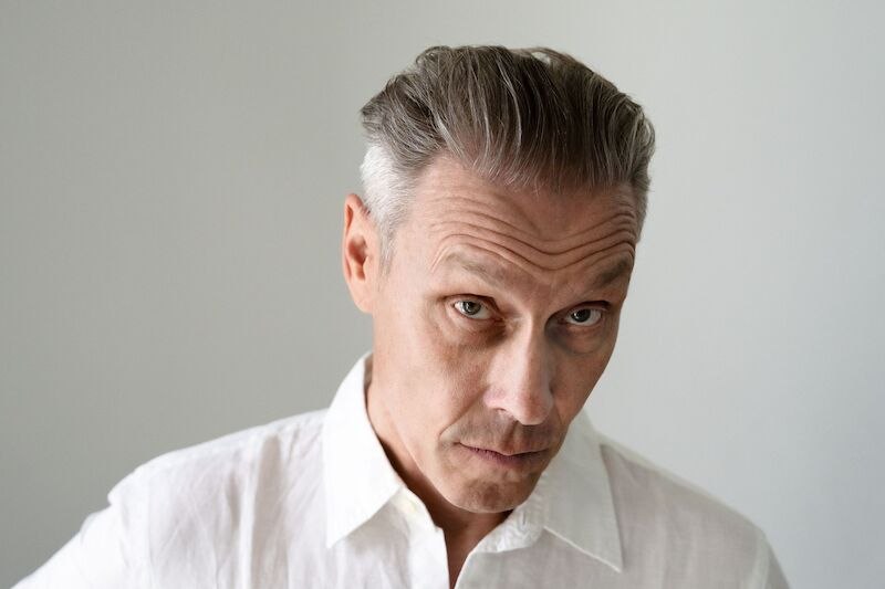 Ein Mann mit grauem Haar und weißem Hemd blickt ernst in die Kamera. Der Hintergrund ist neutral gehalten., © Theaterhaus Stuttgart e.V. Ein Mann mit grauem Haar und weißem Hemd blickt ernst in die Kamera. Der Hintergrund ist neutral gehalten., © Theaterhaus Stuttgart e.V.