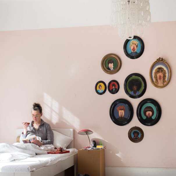 Person sitzt im Bett und liest ein Buch. An der rosa Wand hängen ovale Porträts. Eine Lampe steht auf einem Karton neben dem Bett., © Theater Rampe