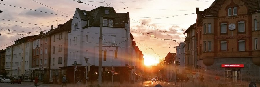 Sonnenuntergang in einer Stadtstraße mit historischen Gebäuden und Straßenbahnkabeln. Menschen überqueren die Straße, während die Sonne zwischen den Häusern untergeht., © SMG Sonnenuntergang in einer Stadtstraße mit historischen Gebäuden und Straßenbahnkabeln. Menschen überqueren die Straße, während die Sonne zwischen den Häusern untergeht., © SMG
