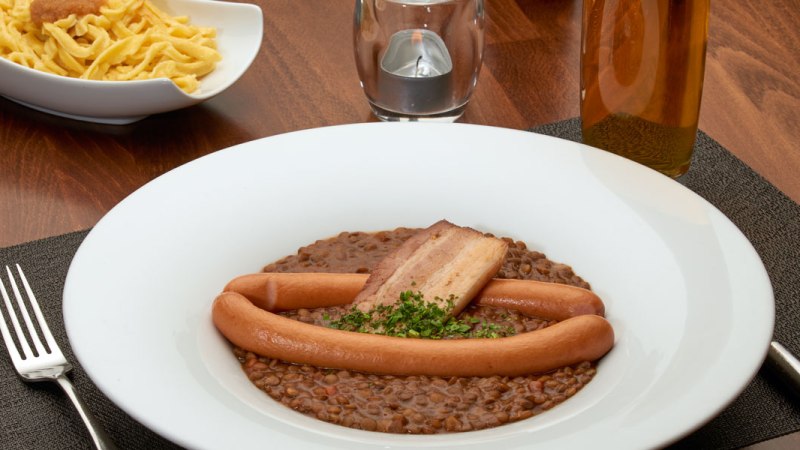 Linseneintopf mit W&uuml;rstchen und Speck in einem wei&szlig;en Teller, daneben eine Sch&uuml;ssel Sp&auml;tzle auf einem Holztisch., &copy; Stuttgart-Marketing GmbH