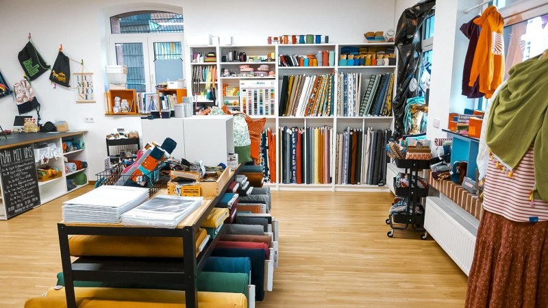 Ein Stoffladen mit Regalen voller bunter Stoffrollen und Nähzubehör. Auf einem Tisch liegen weitere Materialien. An der Wand hängen Taschen., © Lena Beck, Hummelkonig GbR, Stuttgart