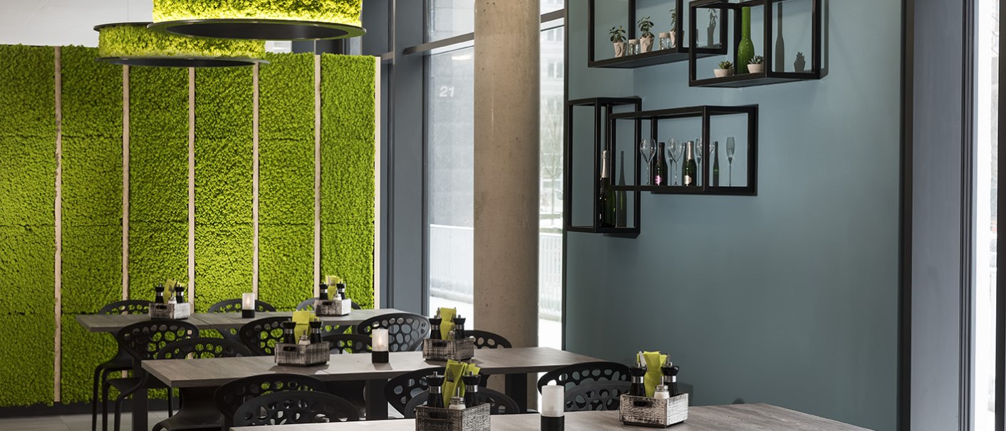 Modernes Restaurant mit grüner Mooswand, stilvollen schwarzen Stühlen und Tischen. An der Wand hängen dekorative Regale mit Flaschen und Pflanzen., © ibis Styles Stuttgart Vaihingen