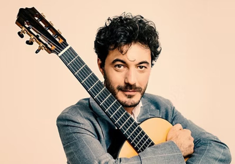 Ein Mann mit dunklem Haar h&auml;lt eine klassische Gitarre und schaut in die Kamera. Er tr&auml;gt einen grauen Anzug vor einem neutralen Hintergrund., &copy; Staatliche Hochschule f&uuml;r Musik und Darstellende Kunst