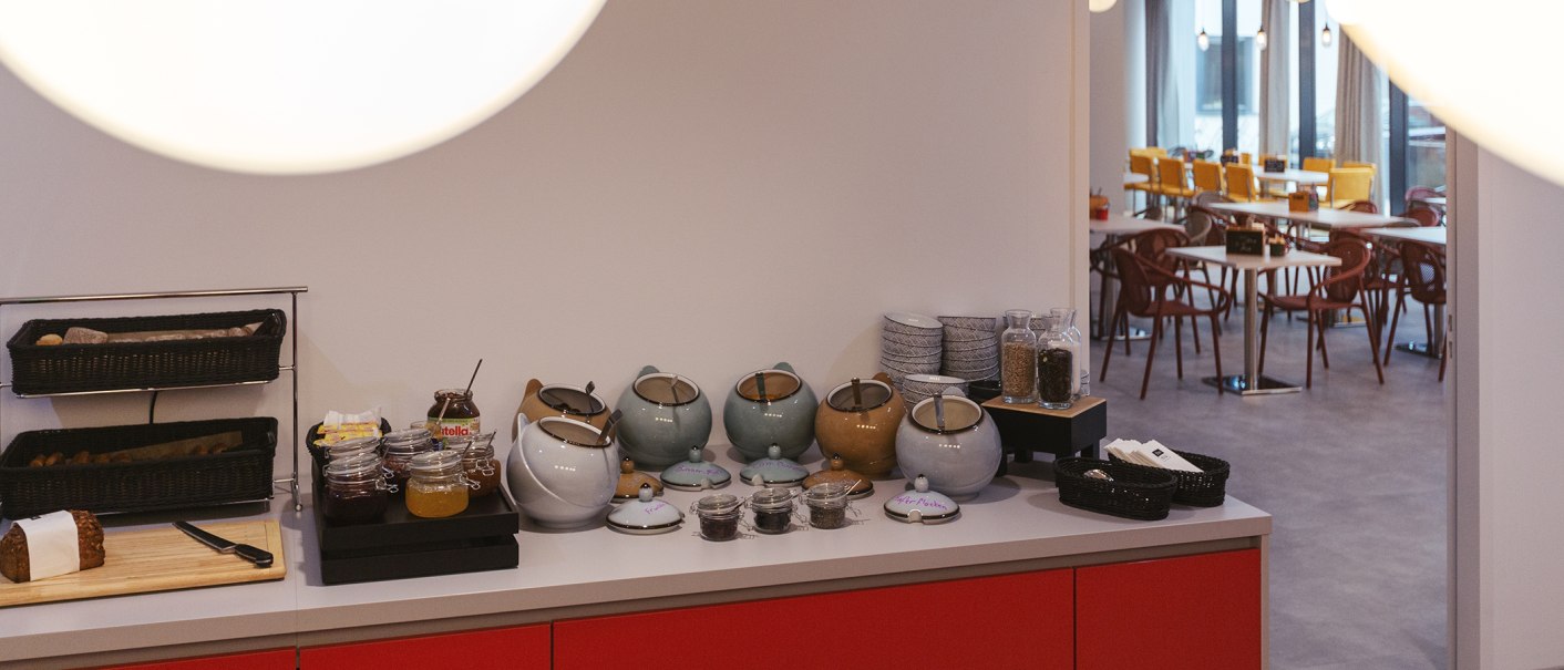 Breakfast buffet with a bread basket, jams, teapots, and tableware. In the background, a dining area with tables and chairs., © the niu Form - Novum Hospitality Breakfast buffet with a bread basket, jams, teapots, and tableware. In the background, a dining area with tables and chairs., © the niu Form - Novum Hospitality